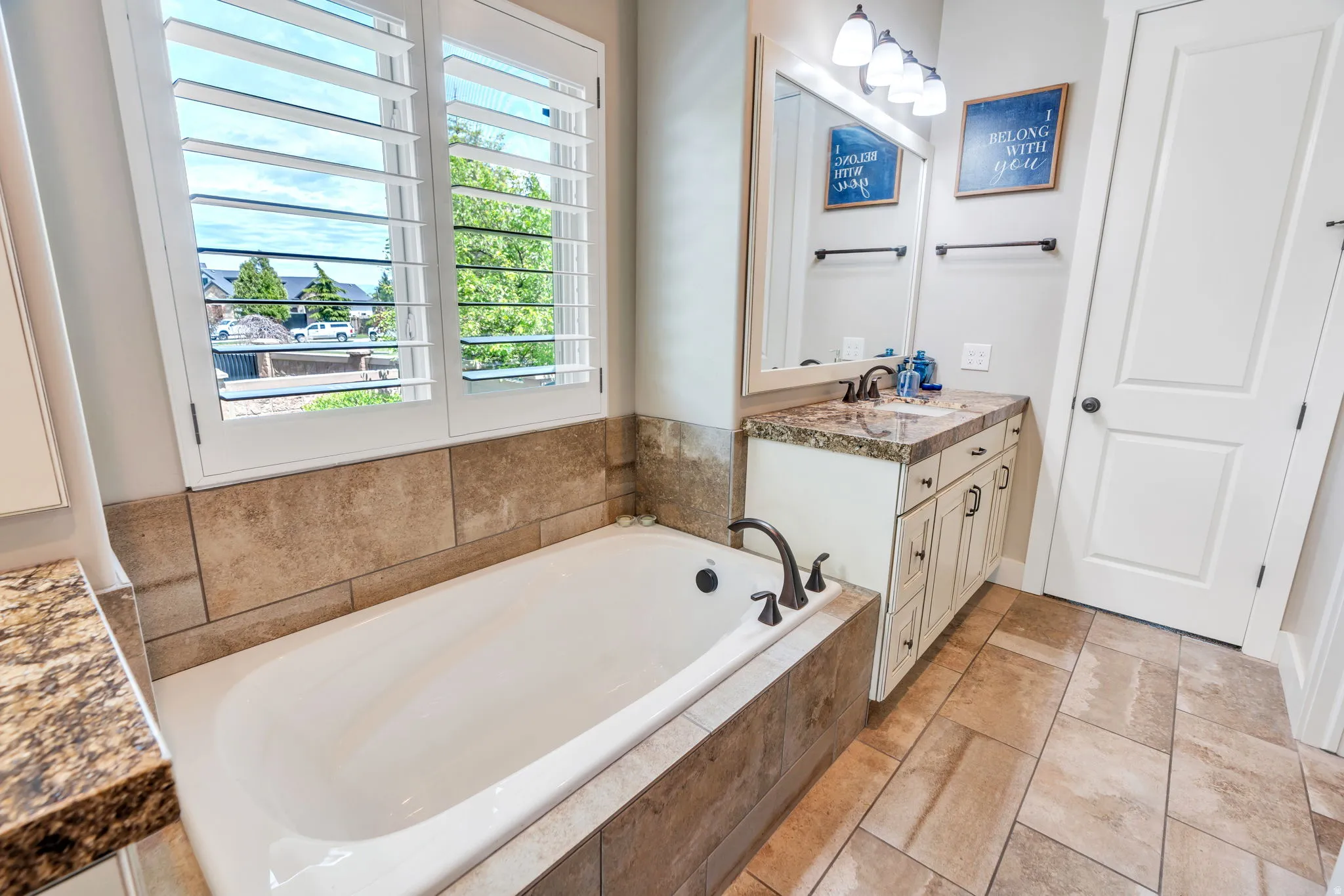 Bathroom featuring a large soaking tub with stone tile surround, plantation shutters, and a vanity with a stone-finish countertop