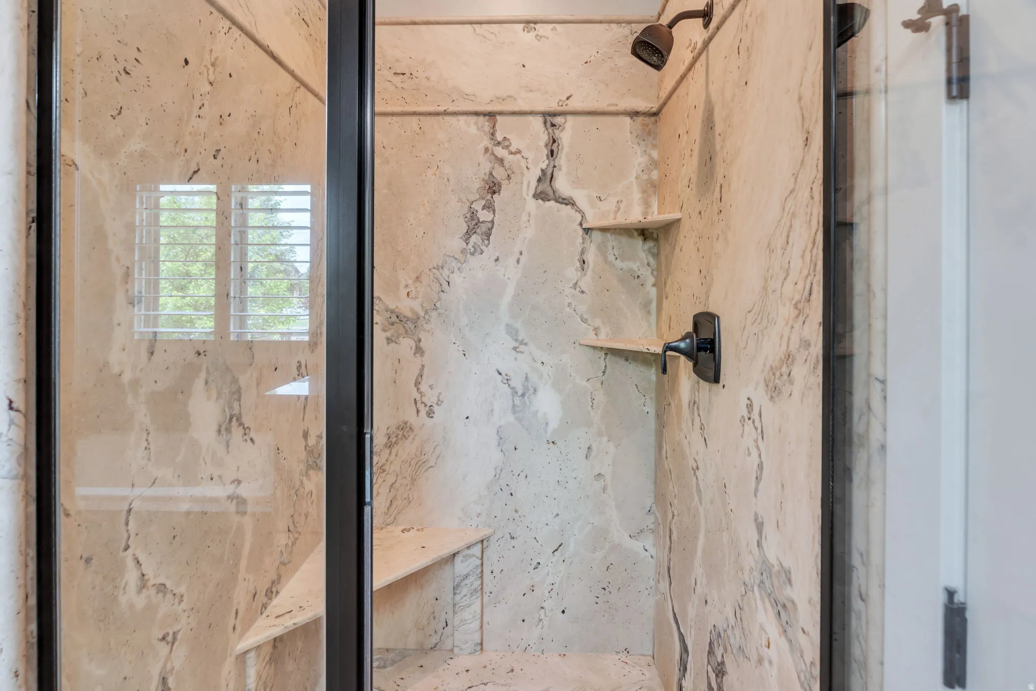 Walk-in shower with natural stone-look wall panels, dark bronze fixtures, built-in corner shelves, and a built-in shower bench