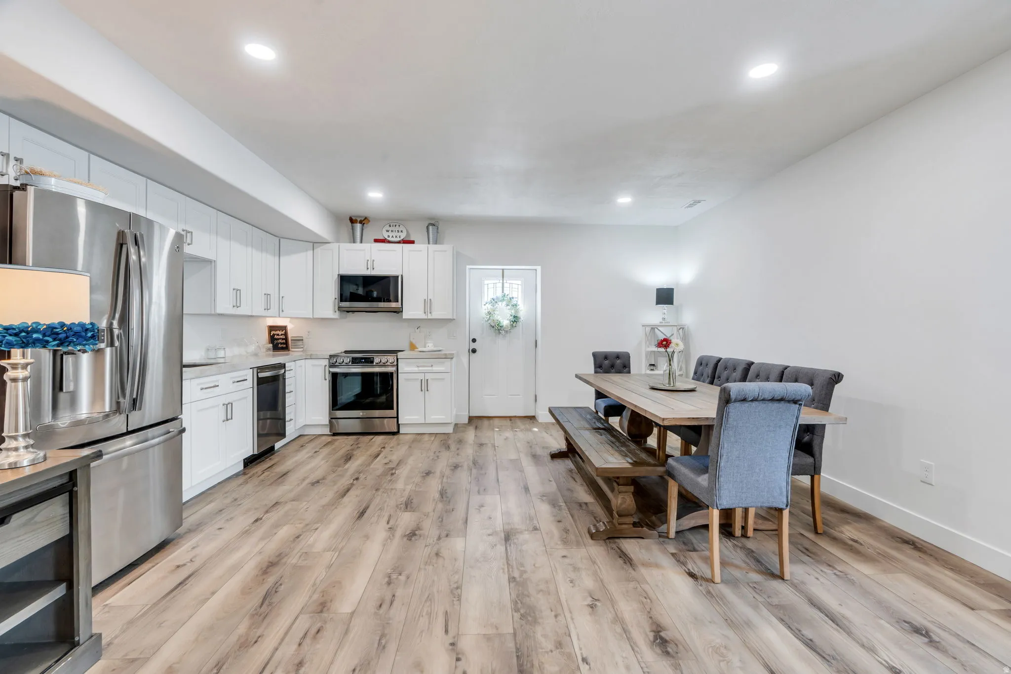 Open-concept living space with wood-finish flooring, recessed lighting, white cabinetry, stainless steel appliances, and a solid white exterior door
