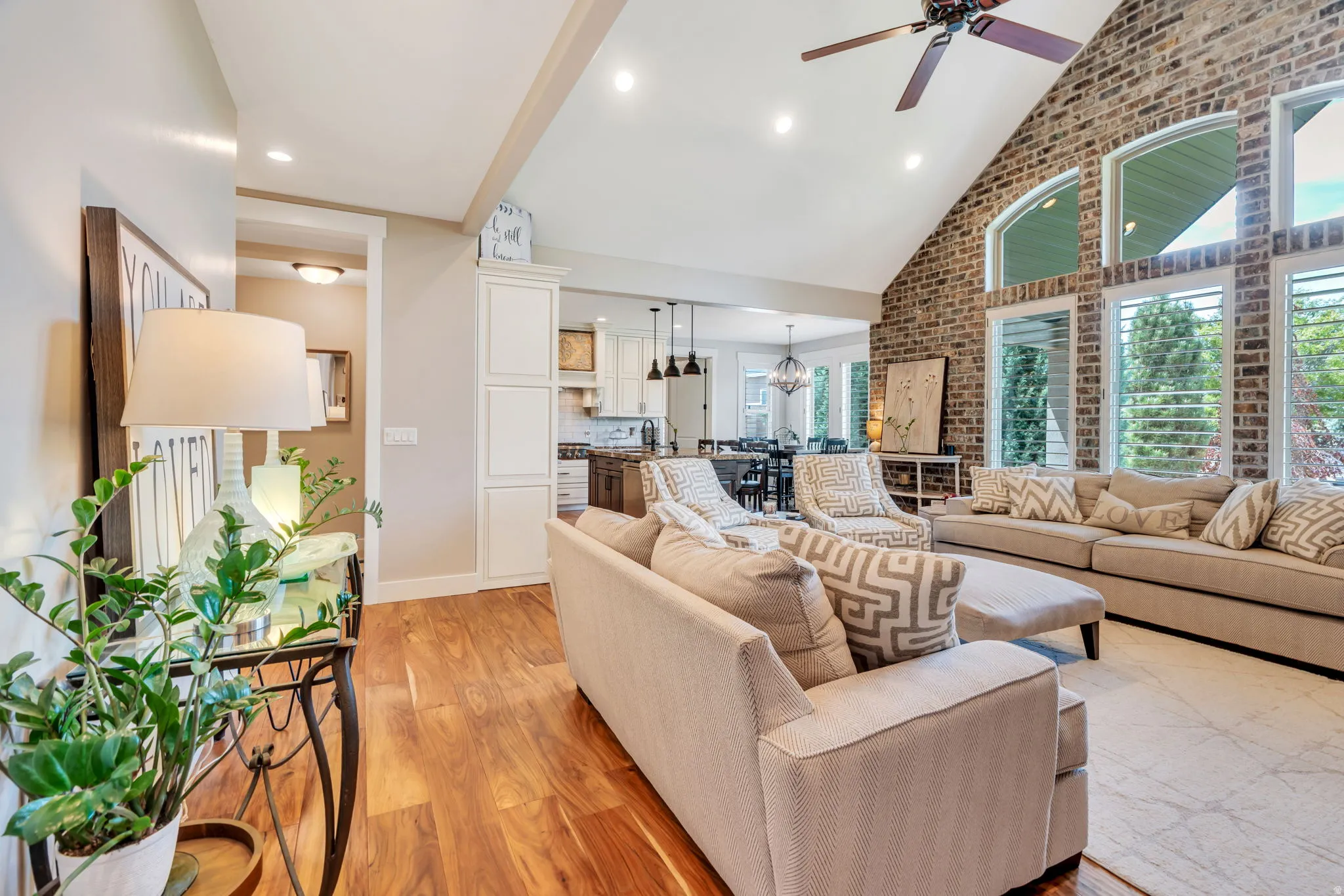 Spacious living area featuring vaulted ceilings, exposed brick accent wall, wood-finish flooring, a ceiling fan, and large windows with plantation shutters