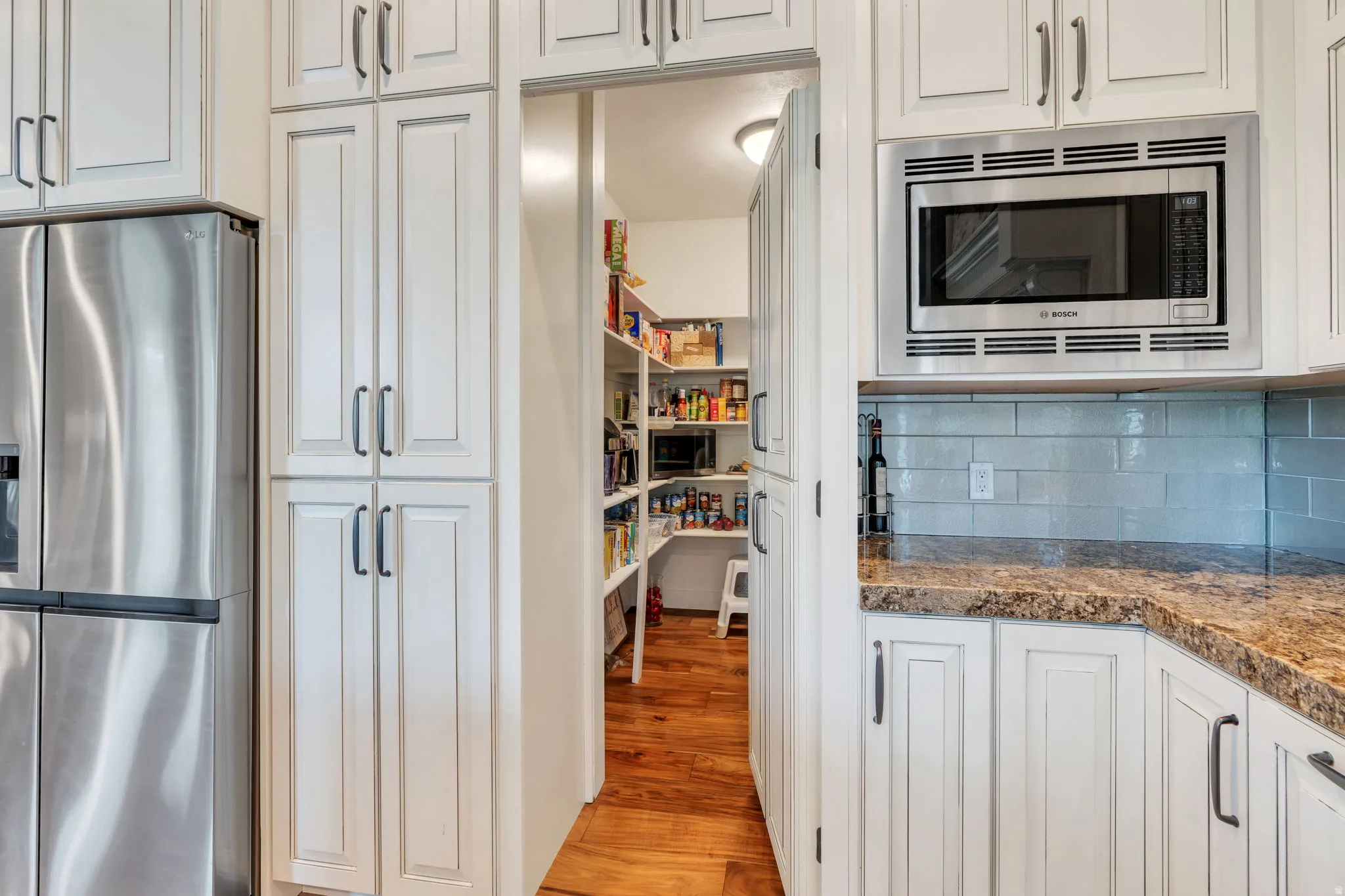 Kitchen pantry with wood-finish flooring, white cabinetry, integrated stainless steel microwave, and light blue subway tile backsplash