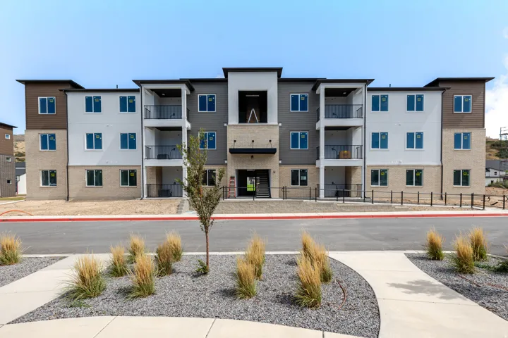 Contemporary multi-story building with a neutral-toned exterior, featuring multiple balconies with black railings, a stone facade on the lower level, and modern landscaping with ornamental grasses and gravel