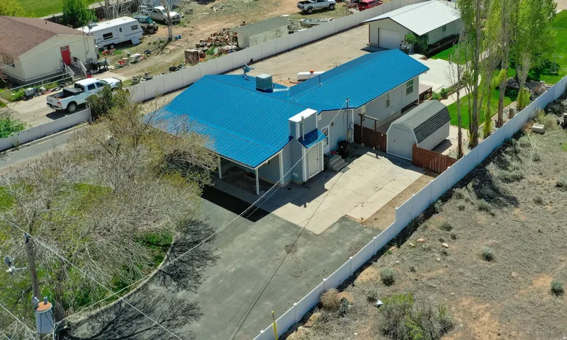 Property featuring a bright blue metal roof, paved driveway, detached storage shed, and white privacy fencing