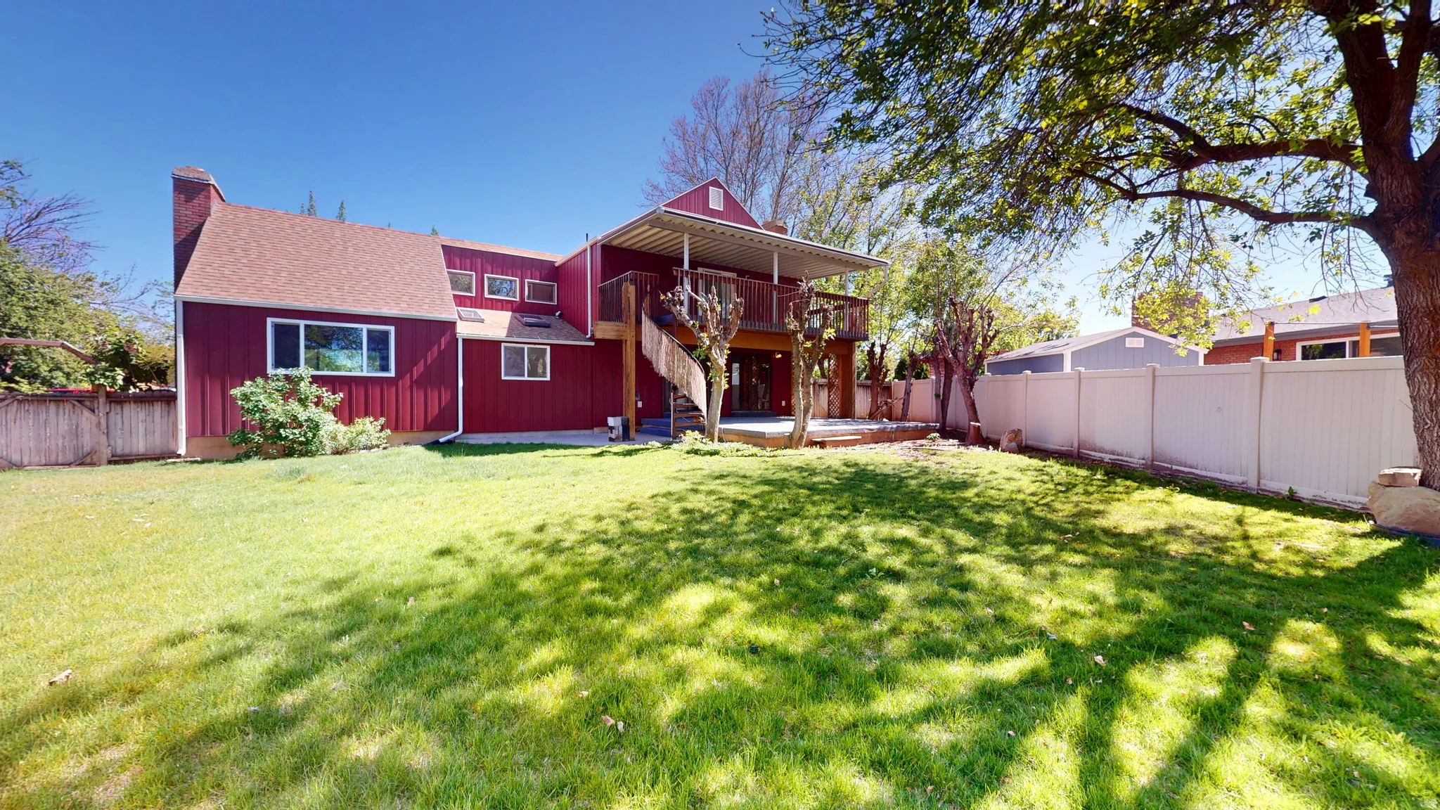 Expansive grassy backyard featuring a multi-level deck, a mature shade tree, privacy fencing, and red siding