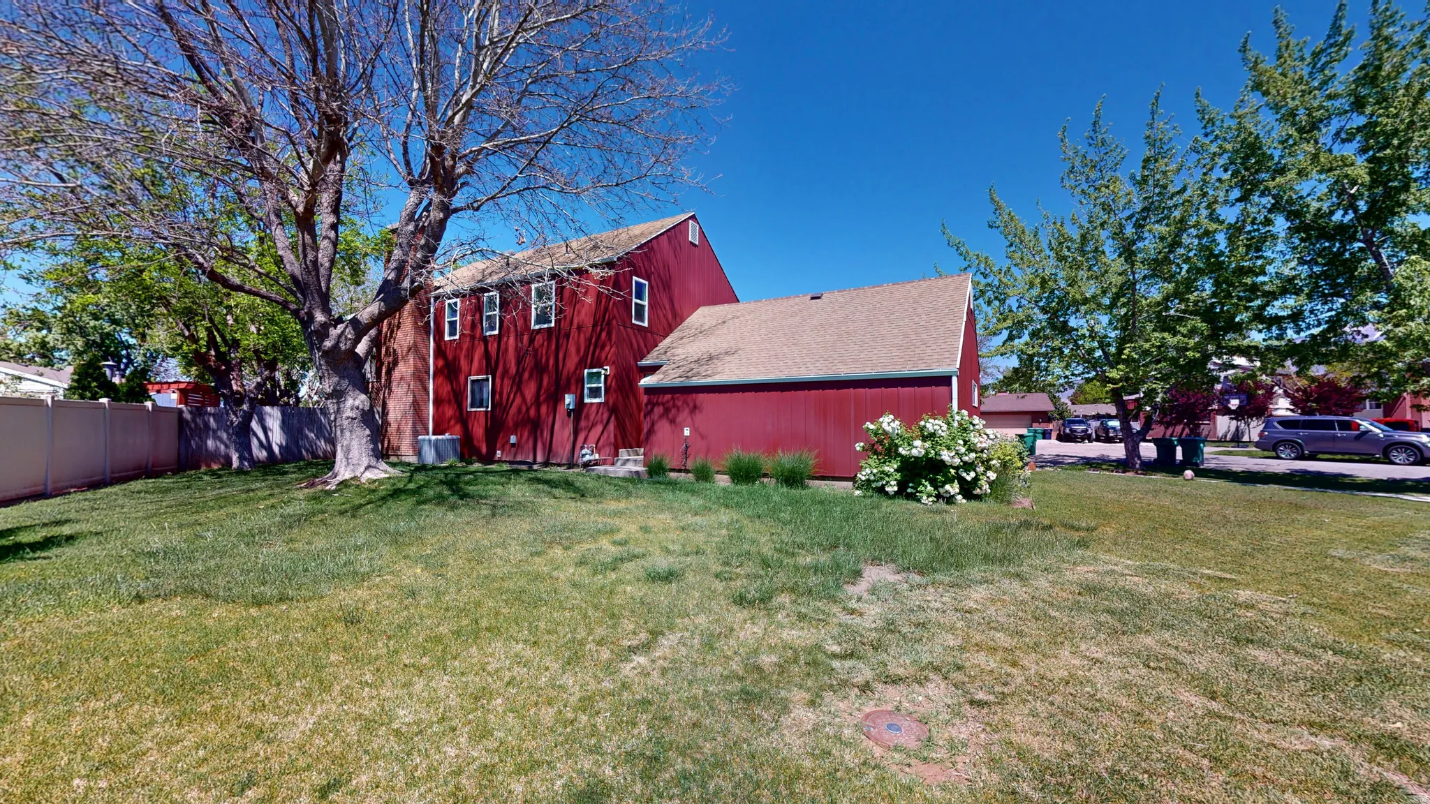 Expansive backyard featuring a mature deciduous tree, a red-sided structure with multiple windows, and an attached red-sided garage with a light-colored roof
