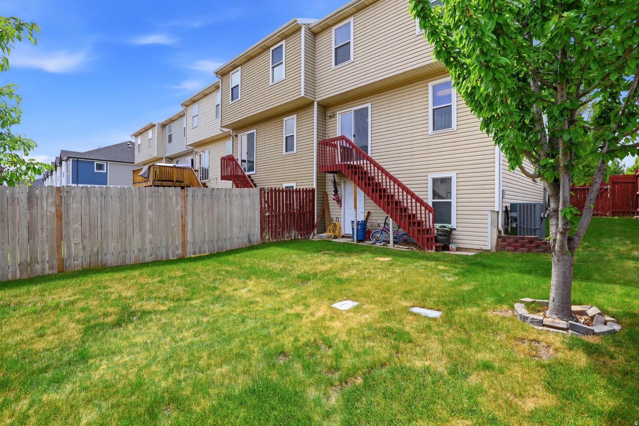 Expansive fenced backyard featuring a mature tree, light beige vinyl siding, red-painted exterior stairs, and a ground-level patio space