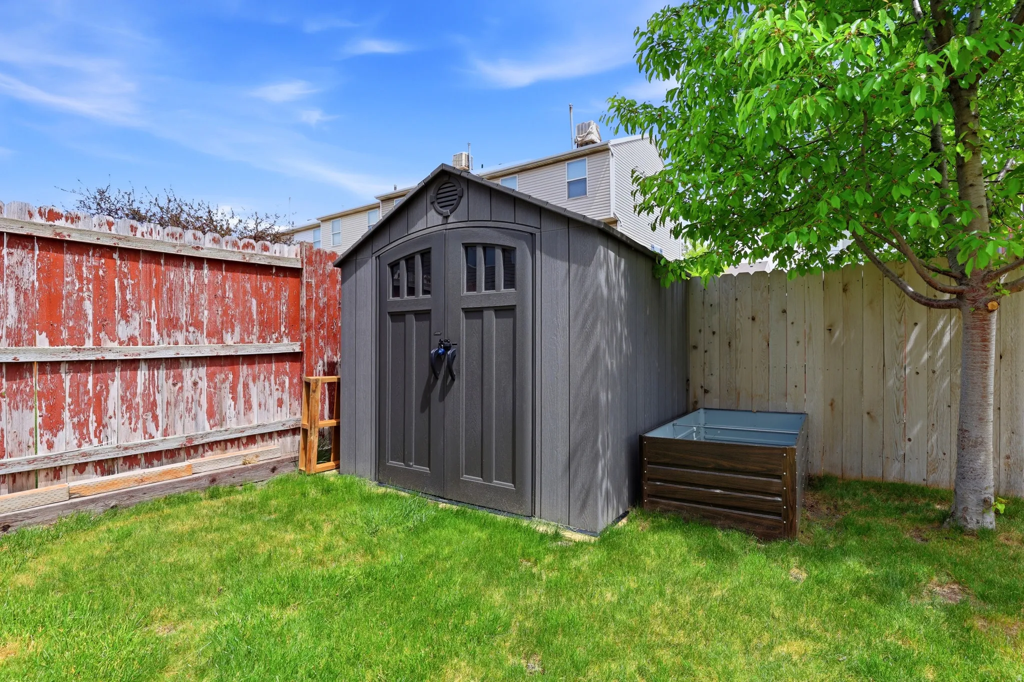 Backyard area featuring a dark gray storage shed with arched double doors, a wood-slat planter box, and a mature tree