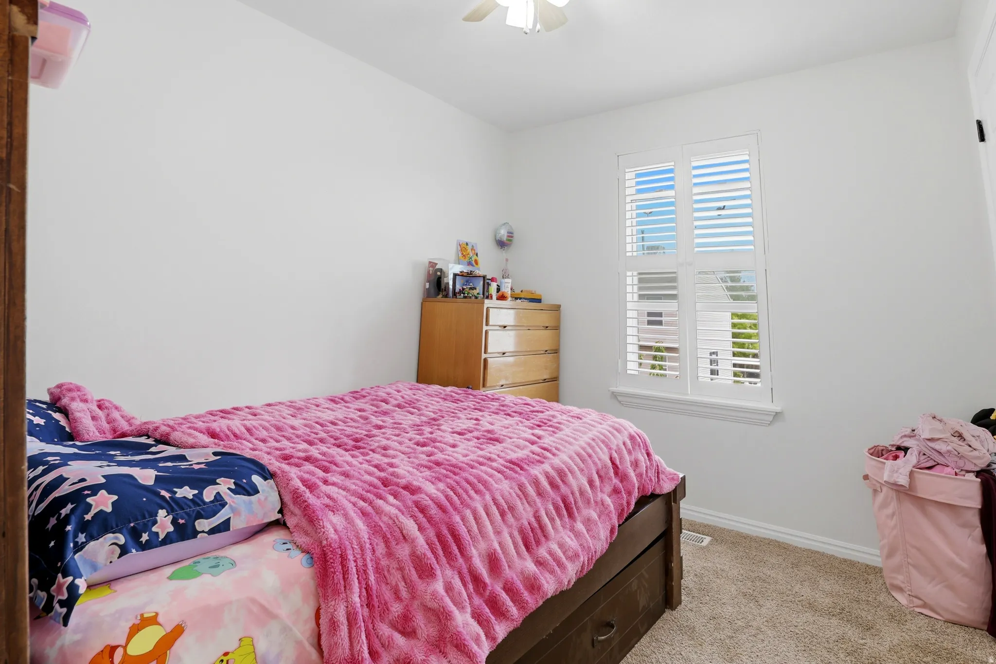 Bright room with white walls, carpet flooring, a window with white plantation shutters, and a ceiling fan with integrated lighting