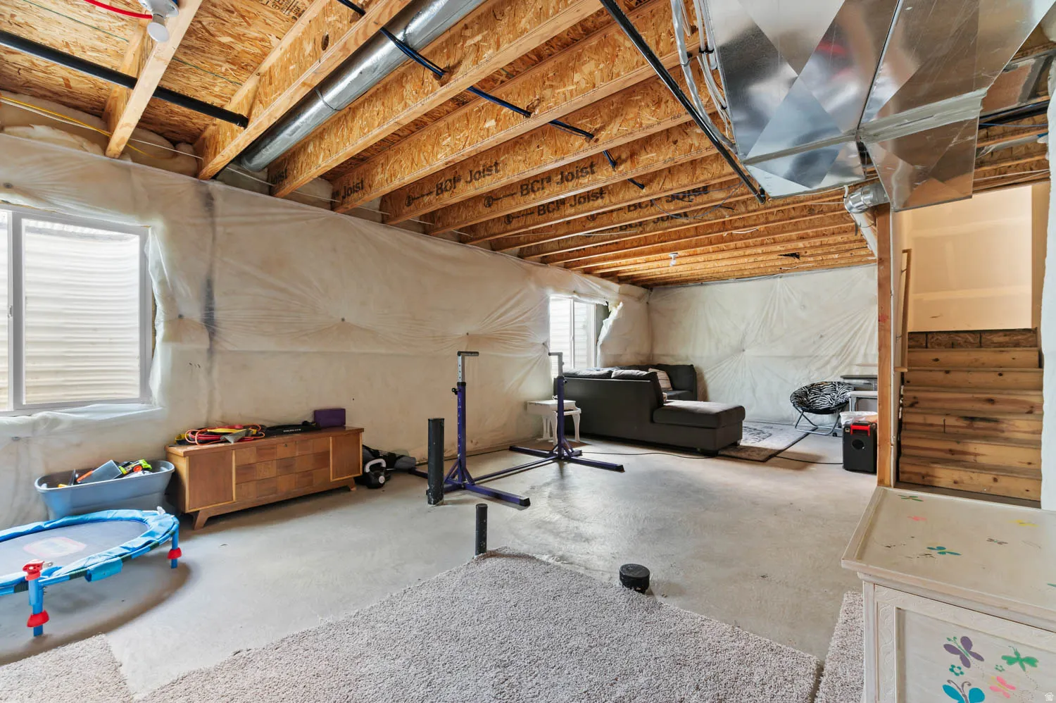 Unfinished basement space featuring exposed wood joists, visible ductwork, concrete flooring, and a window with horizontal blinds