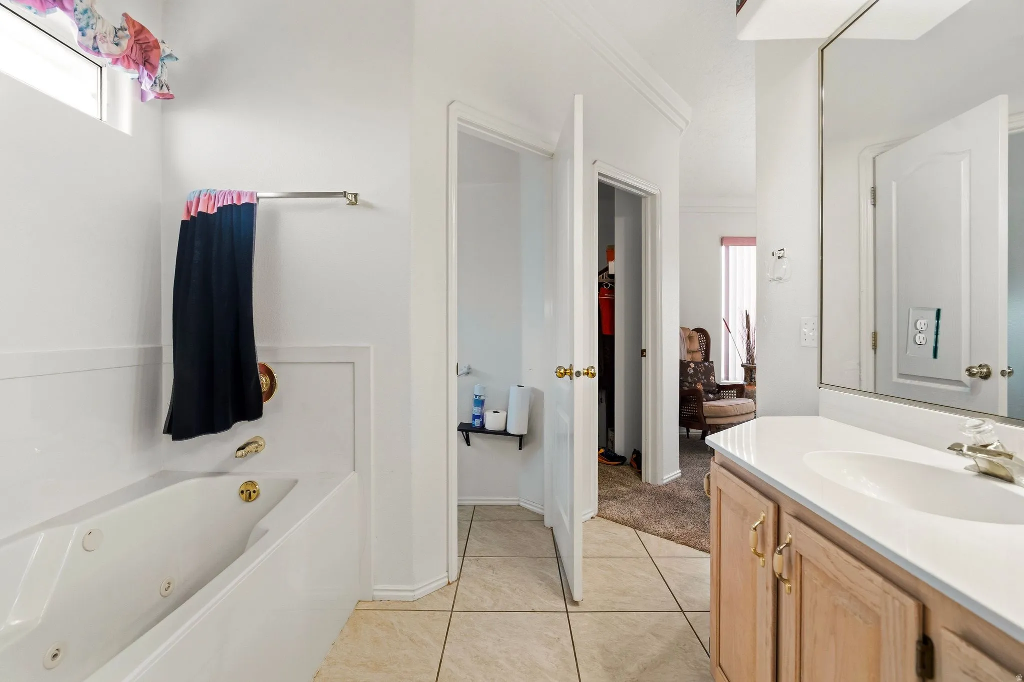 Bathroom featuring a built-in tub with gold-tone fixtures, tile flooring, a single vanity with wood-finish cabinetry, a large wall-mounted mirror, and a transom window