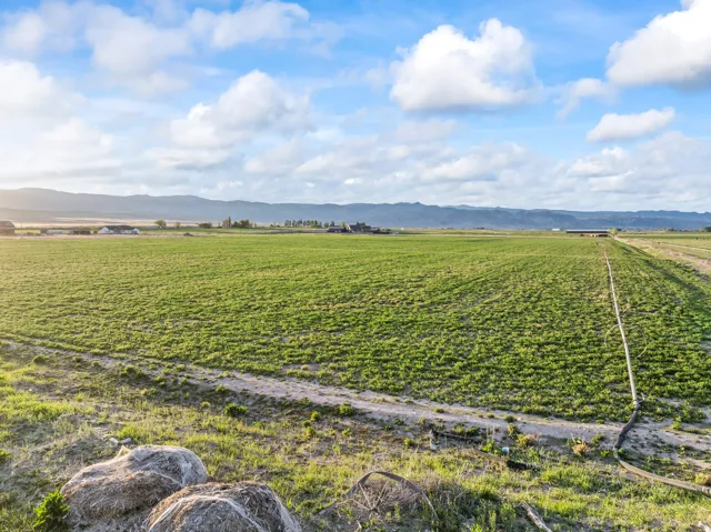 Expansive agricultural land featuring verdant fields, distant mountain ranges, a clear blue sky with scattered clouds, and visible irrigation infrastructure