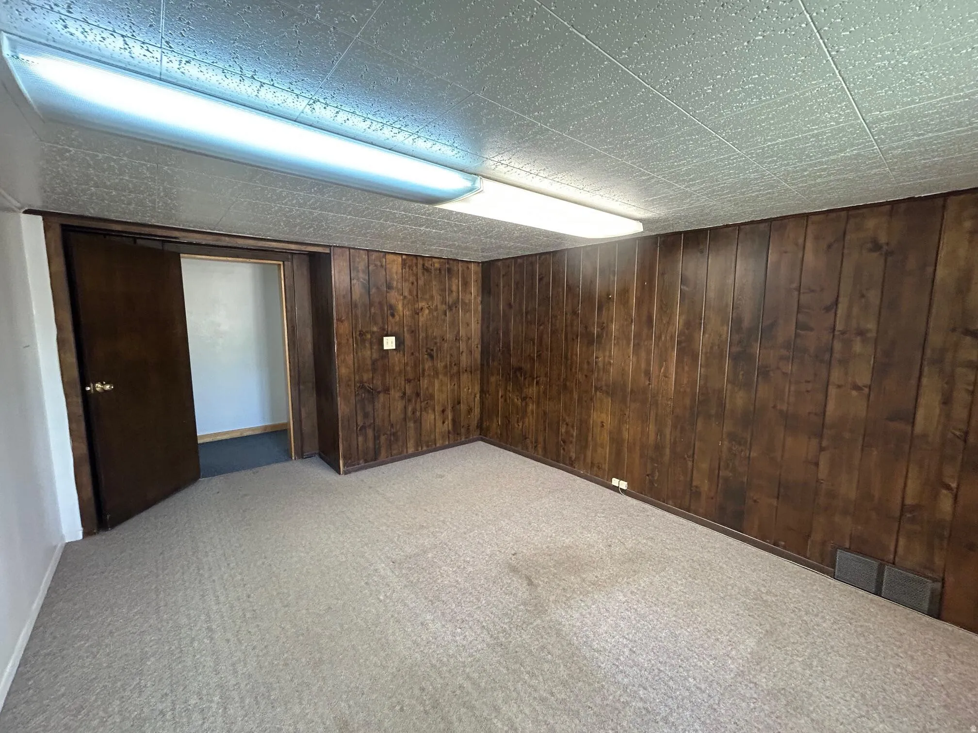 Carpeted room featuring wood-panel walls, a drop ceiling with integrated fluorescent lighting, and a dark wood-finish door