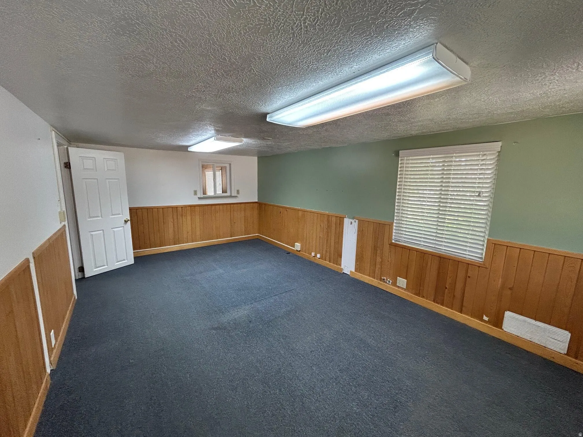 Spacious interior featuring wood-panel wainscoting, textured ceiling, two fluorescent light fixtures, and a window with blinds