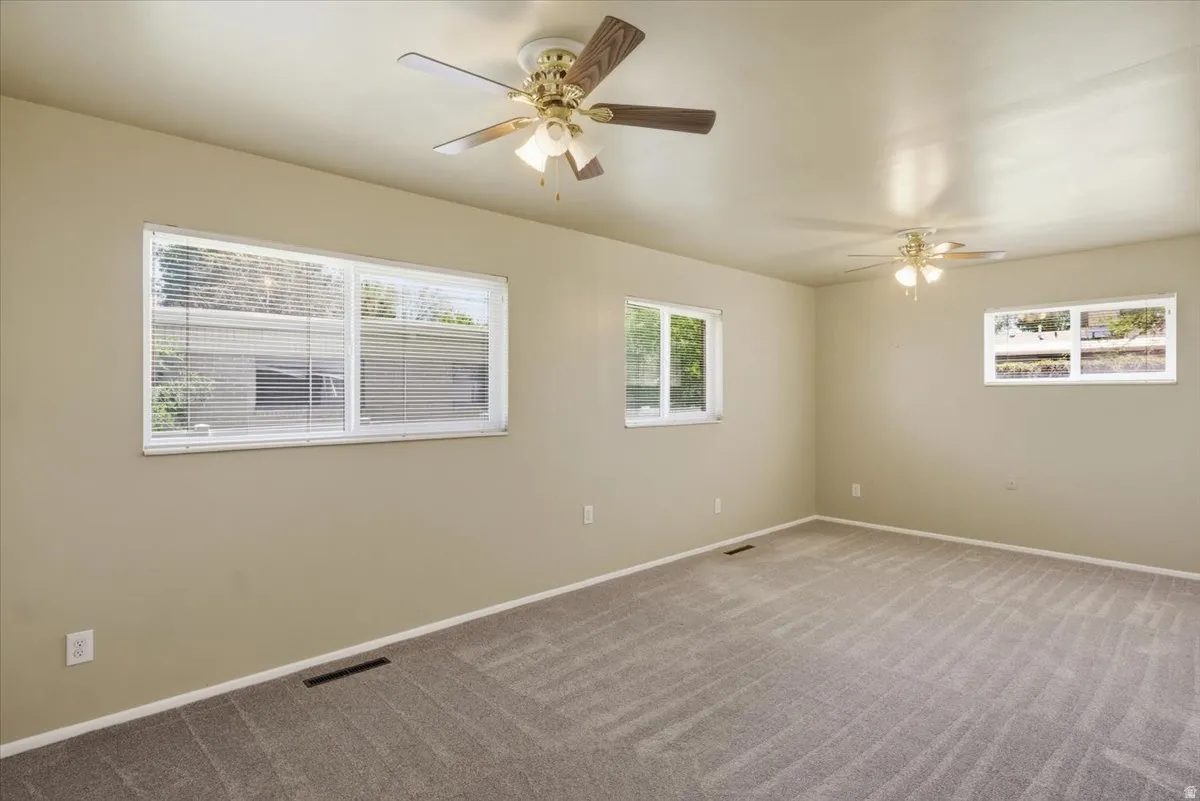 Spacious room featuring neutral-toned walls, ceiling fans with integrated lighting, multiple windows with blinds, and patterned carpet flooring