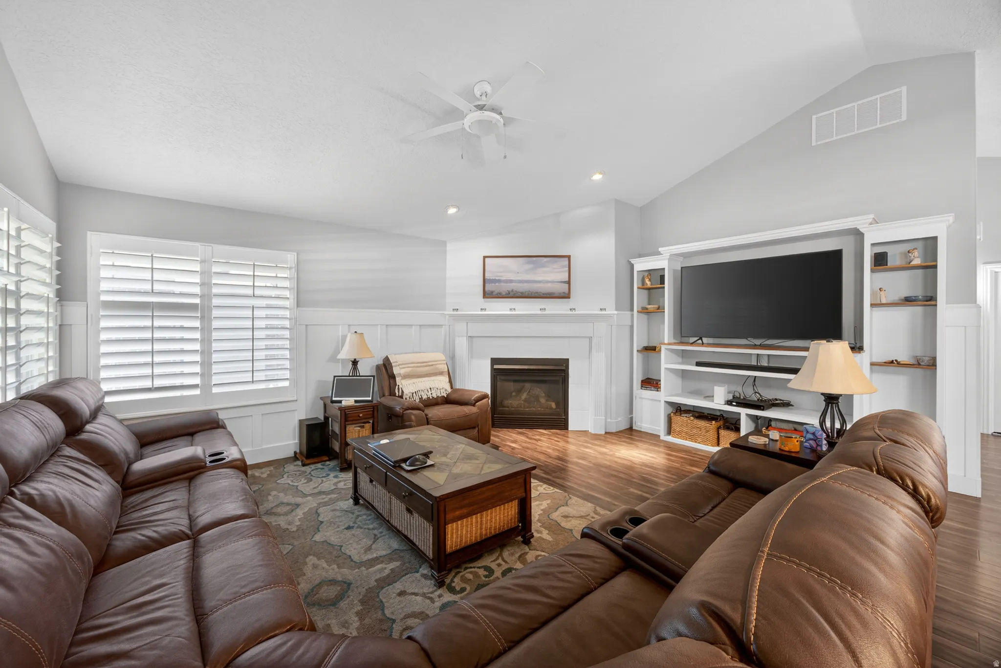 Living area featuring wood-finish flooring, a vaulted ceiling, a gas fireplace with a white mantel, custom built-in shelving, and plantation shutters