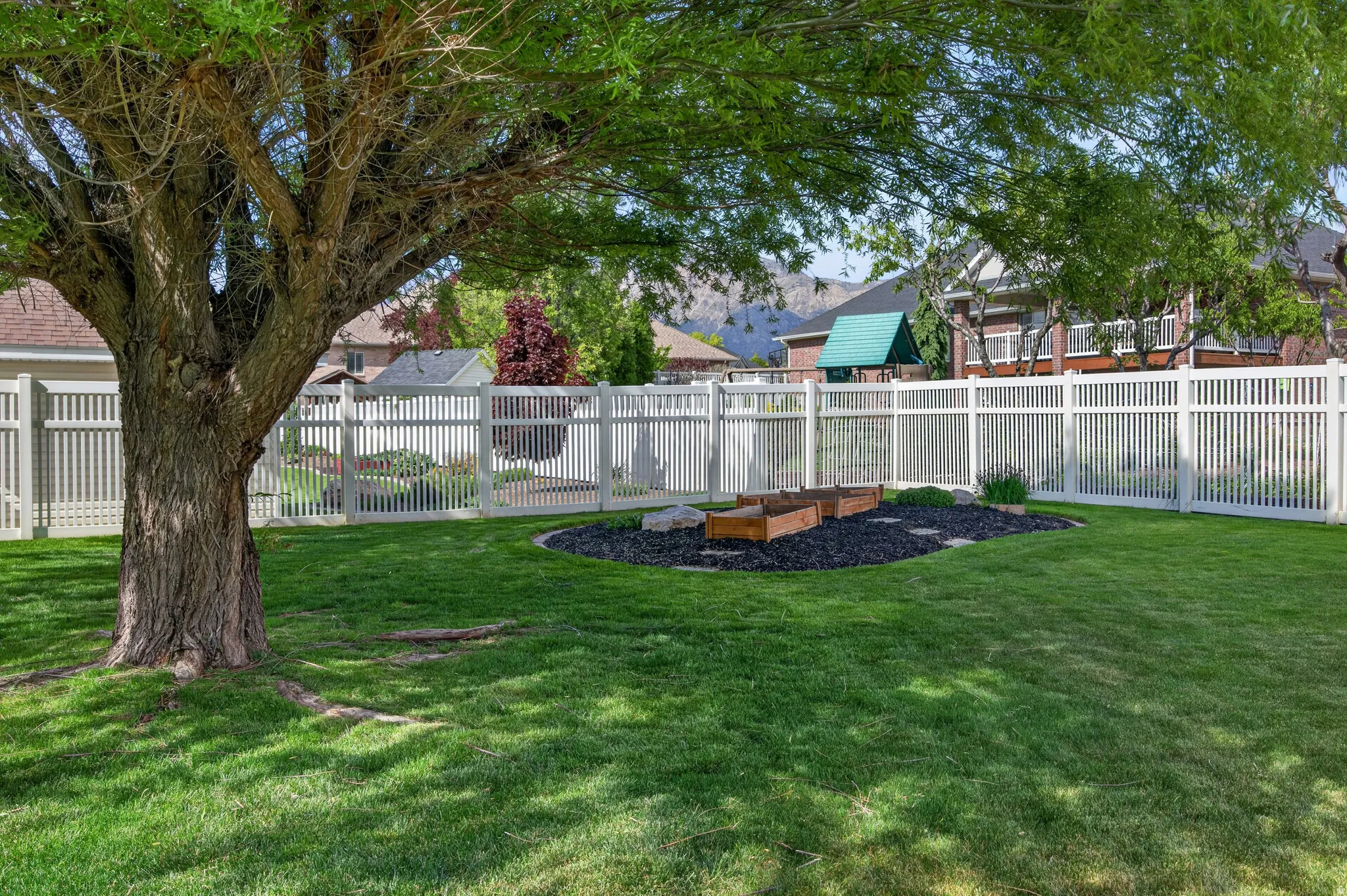 Expansive green lawn with a mature shade tree, white picket fencing, and a landscaped garden bed featuring wood planters