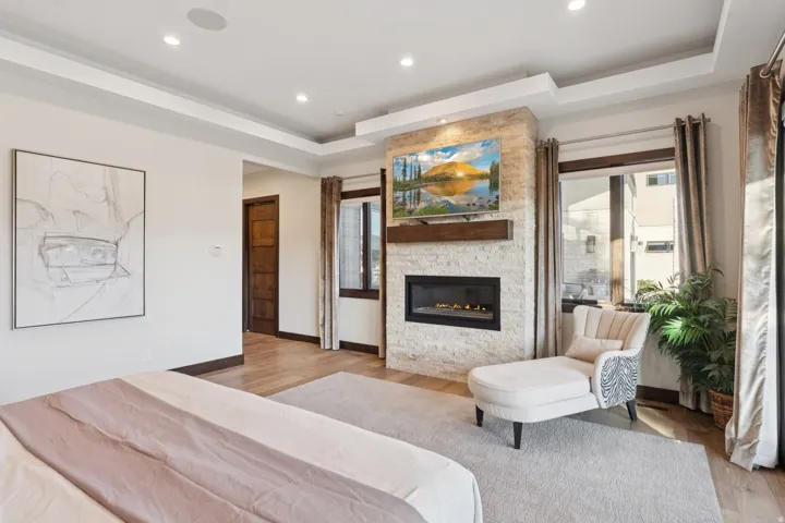 Spacious room featuring a stone-clad fireplace with a built-in television, tray ceiling with recessed lighting, wood-finish flooring, and a solid wood paneled door