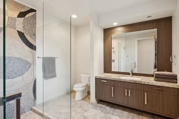 Glass-enclosed shower with a decorative mosaic tile wall, a contemporary wood-finish vanity with an integrated sink, a large mirror, and recessed lighting