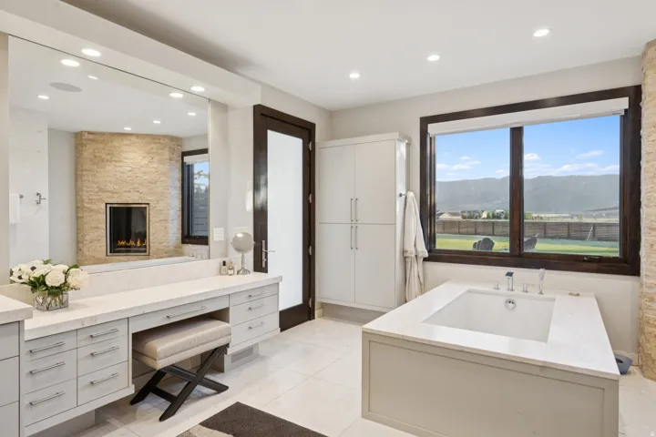 Spacious bathroom featuring a built-in soaking tub, stone-clad fireplace, integrated vanity with ample storage, large mirror, and panoramic mountain scenery