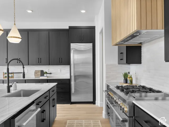 Gourmet kitchen with black cabinetry, stainless steel appliances, large kitchen island with integrated sink, wood-finish flooring, and a decorative wood-slat range hood