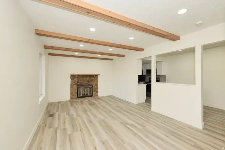 Living area featuring wood-finish flooring, exposed ceiling beams, recessed lighting, a stone fireplace with a wood mantle, and a pass-through window to the kitchen