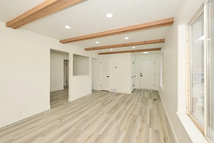 Spacious room featuring wood-finish flooring, exposed wood ceiling beams, recessed lighting, white walls, and a large window