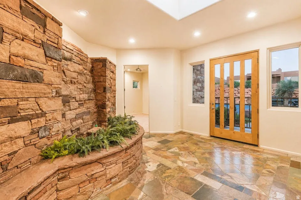 Stone accent wall, multi-tone slate flooring, recessed lighting, wood-paneled door with glass inserts, and multiple windows