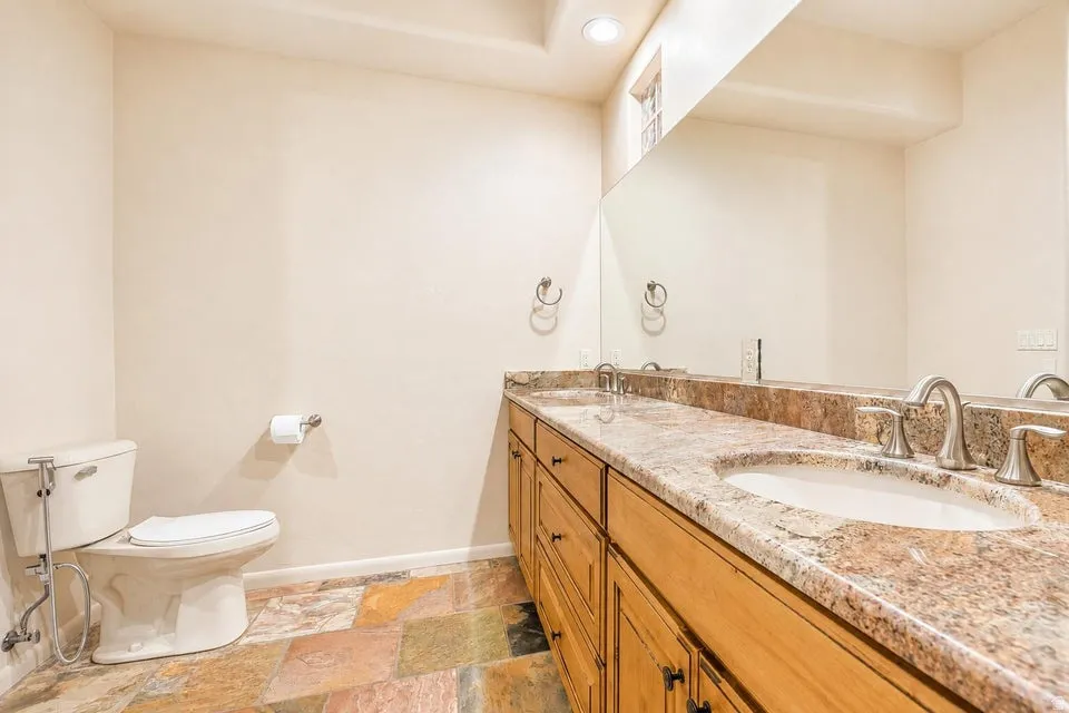 Dual vanity with granite countertop, integrated sinks, wood-finish cabinetry, and multi-toned stone flooring