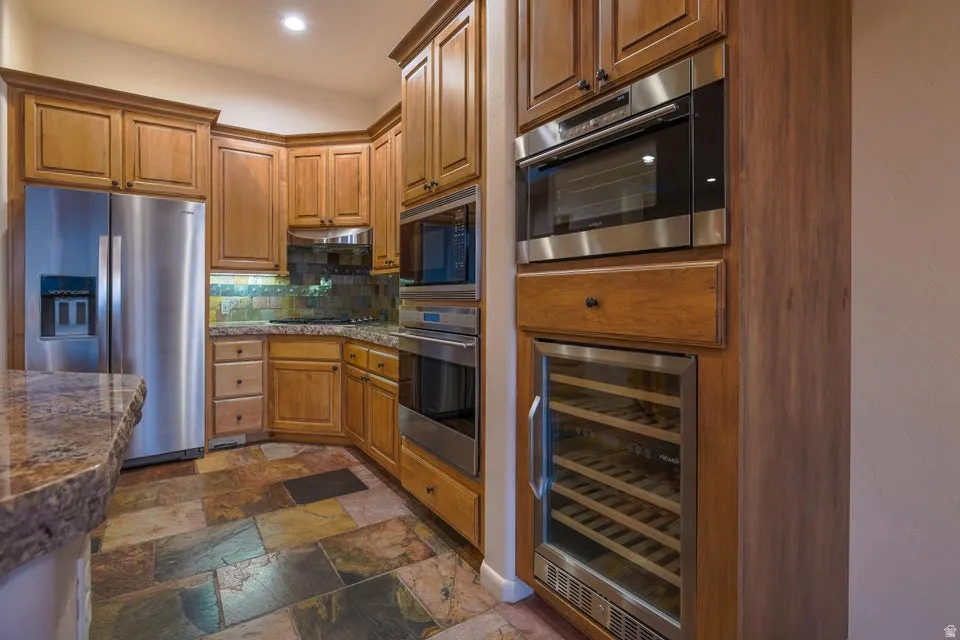 Wood cabinetry with crown molding, stainless steel appliances, stone slab countertops, tiled backsplash, and multi-tone tile flooring