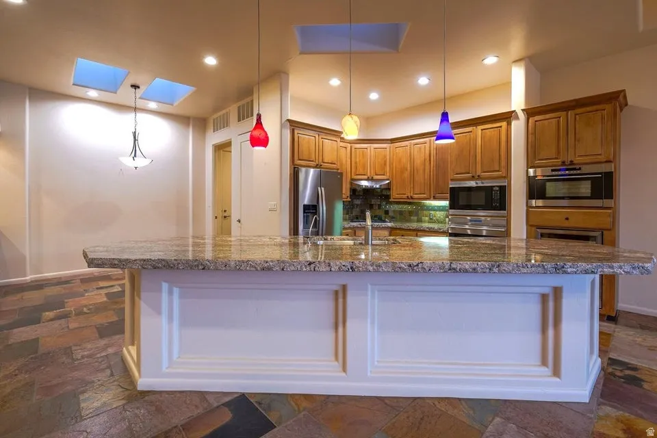 Expansive kitchen island with a stone countertop, built-in stainless steel appliances, wood cabinetry, and slate-style flooring