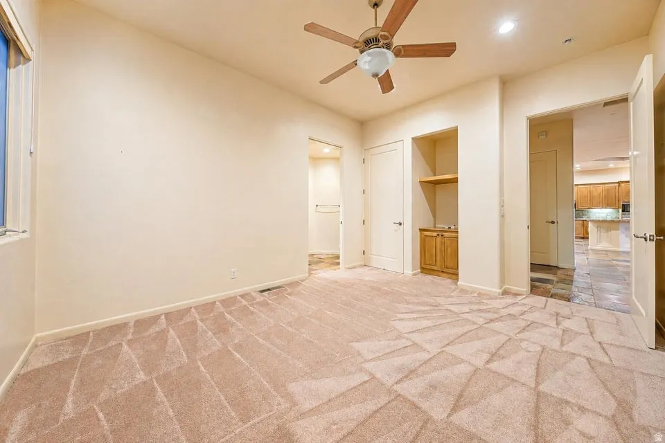 Neutral tone walls, patterned carpeting, ceiling fan with integrated lighting, built-in wooden cabinetry with shelving, and tile flooring in the adjacent space