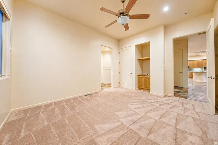 Neutral tone walls, patterned carpeting, ceiling fan with integrated lighting, built-in wooden cabinetry with shelving, and tile flooring in the adjacent space