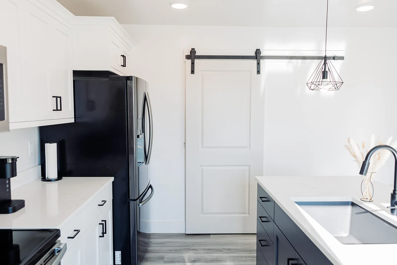 Modern kitchen featuring a white barn door, black refrigerator, white cabinetry with dark hardware, a kitchen island with an undermount sink, and wood-finish flooring