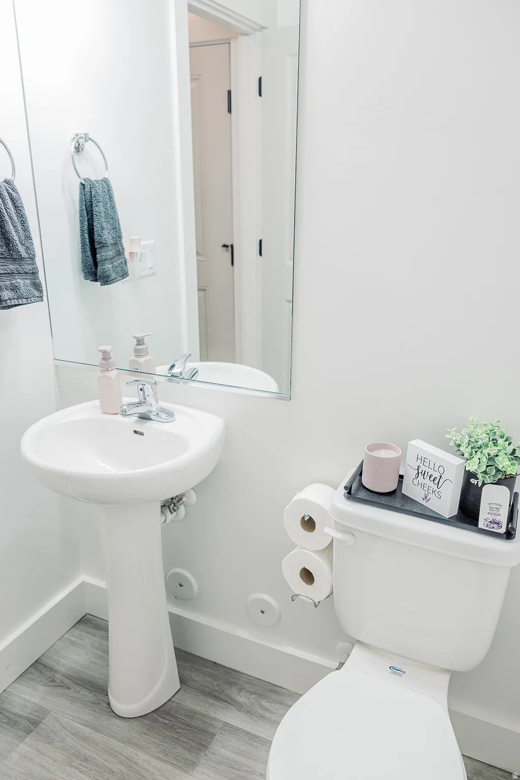 Pedestal sink with chrome faucet, large wall mirror, and grey wood-finish flooring