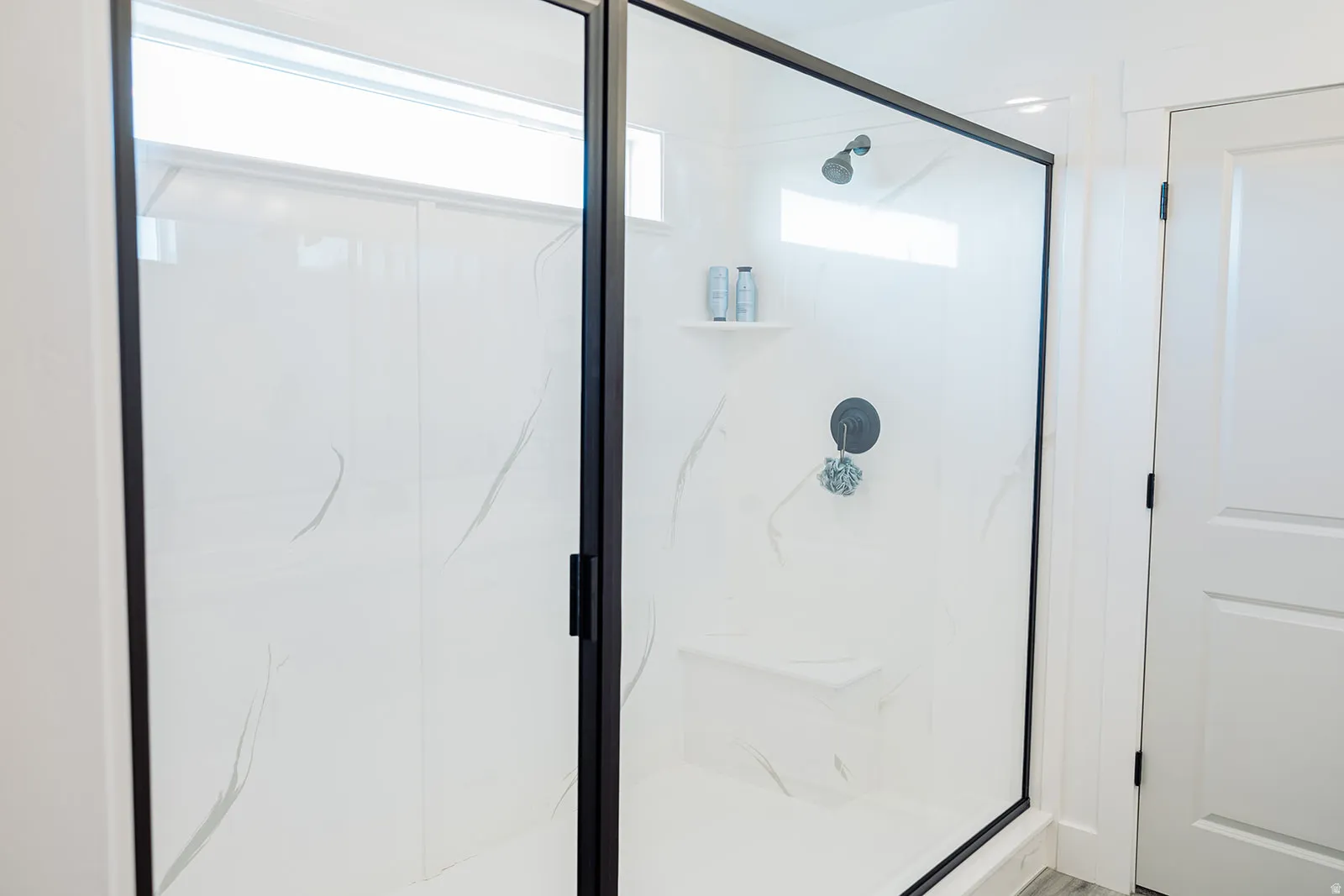 Spacious shower enclosure featuring a black-framed glass door, white marbled wall panels, matte black shower fixtures, an integrated shower bench, and a transom window for natural light