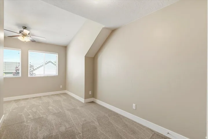 Vaulted ceiling room featuring two windows with horizontal blinds, a ceiling fan with integrated lighting, neutral-toned walls, and plush carpet flooring