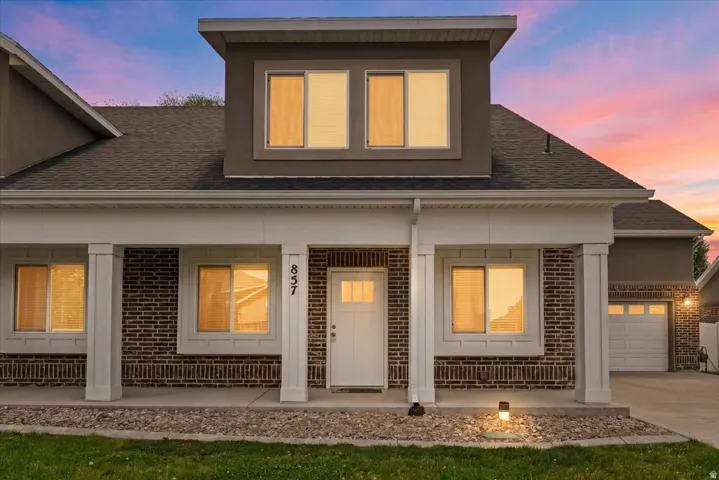 Brick and stucco exterior featuring a covered porch with white columns, a prominent second-story dormer with multiple windows, an attached garage, and a manicured lawn