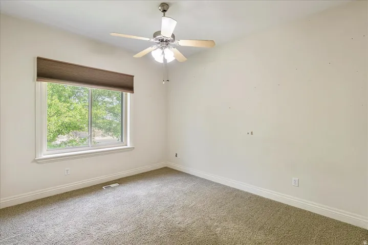 Spacious room featuring a large window with exterior landscaping, neutral wall tones, a ceiling fan with integrated lighting, and plush carpet flooring