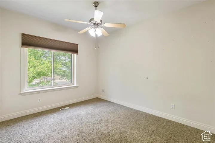 Spacious room featuring a large window with exterior landscaping, neutral wall tones, a ceiling fan with integrated lighting, and plush carpet flooring