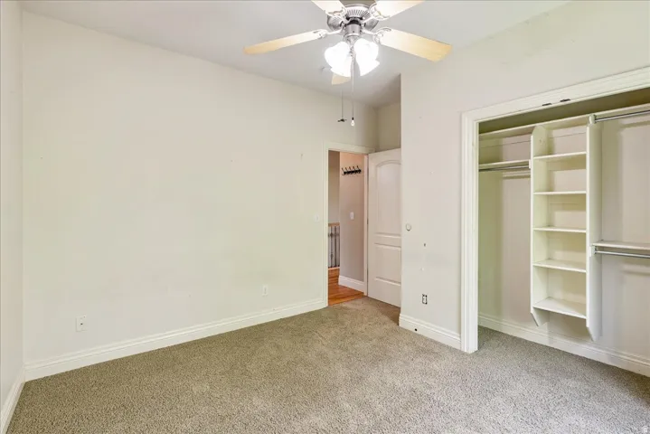 Room featuring neutral carpeting, a ceiling fan with light fixture, and a built-in closet organization system with shelving and hanging rods