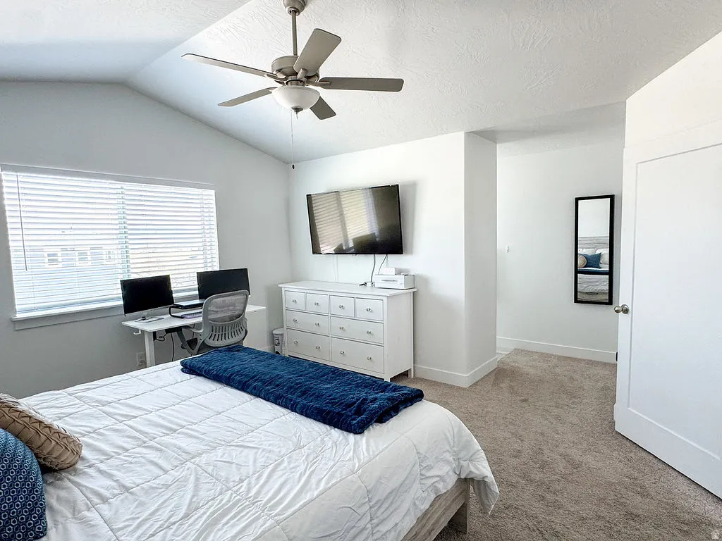 Room featuring an angled vaulted ceiling with a flush-mount ceiling fan, a large window with horizontal blinds, neutral carpet flooring, and a flat-panel door