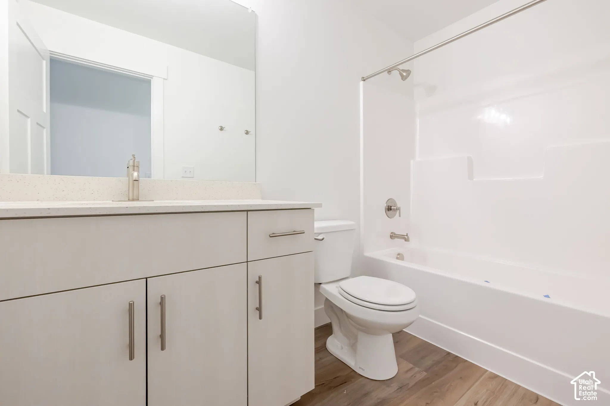 White vanity with a light-toned countertop, a rectangular mirror, a white toilet, a white shower-over-tub unit, and wood-finish flooring