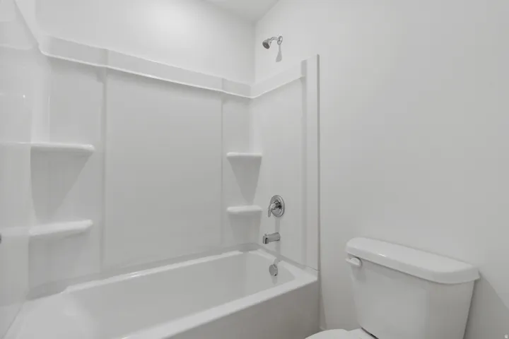 White bathtub with an integrated shower surround featuring built-in shelving, chrome fixtures, and a coordinating toilet