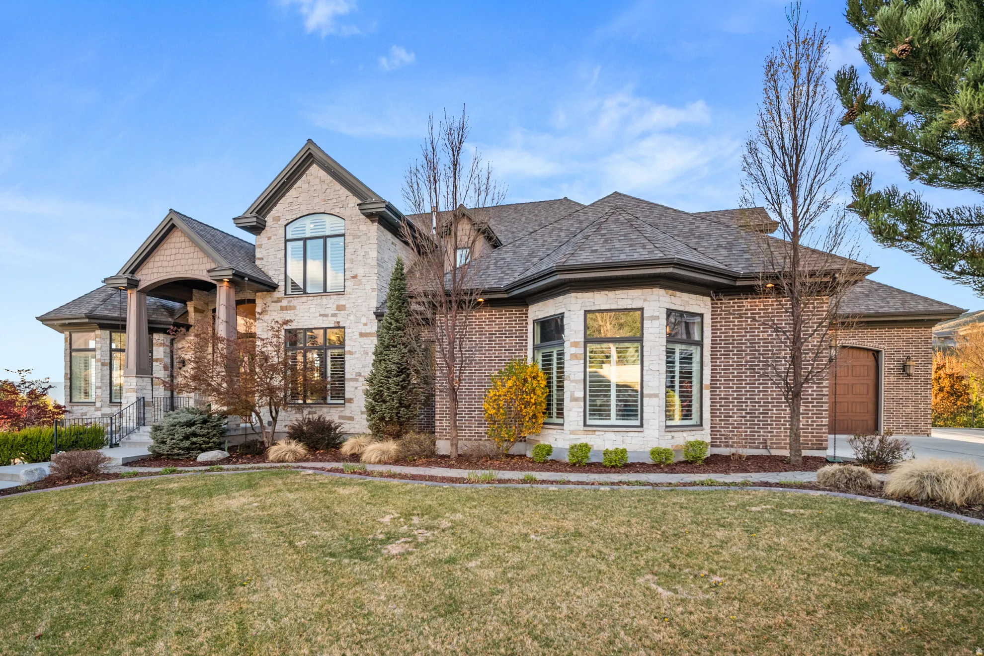 Stone and brick exterior featuring a prominent arched entry, multiple gables, a bay window with plantation shutters, a wood-finish garage door, and a manicured front lawn with mature landscaping