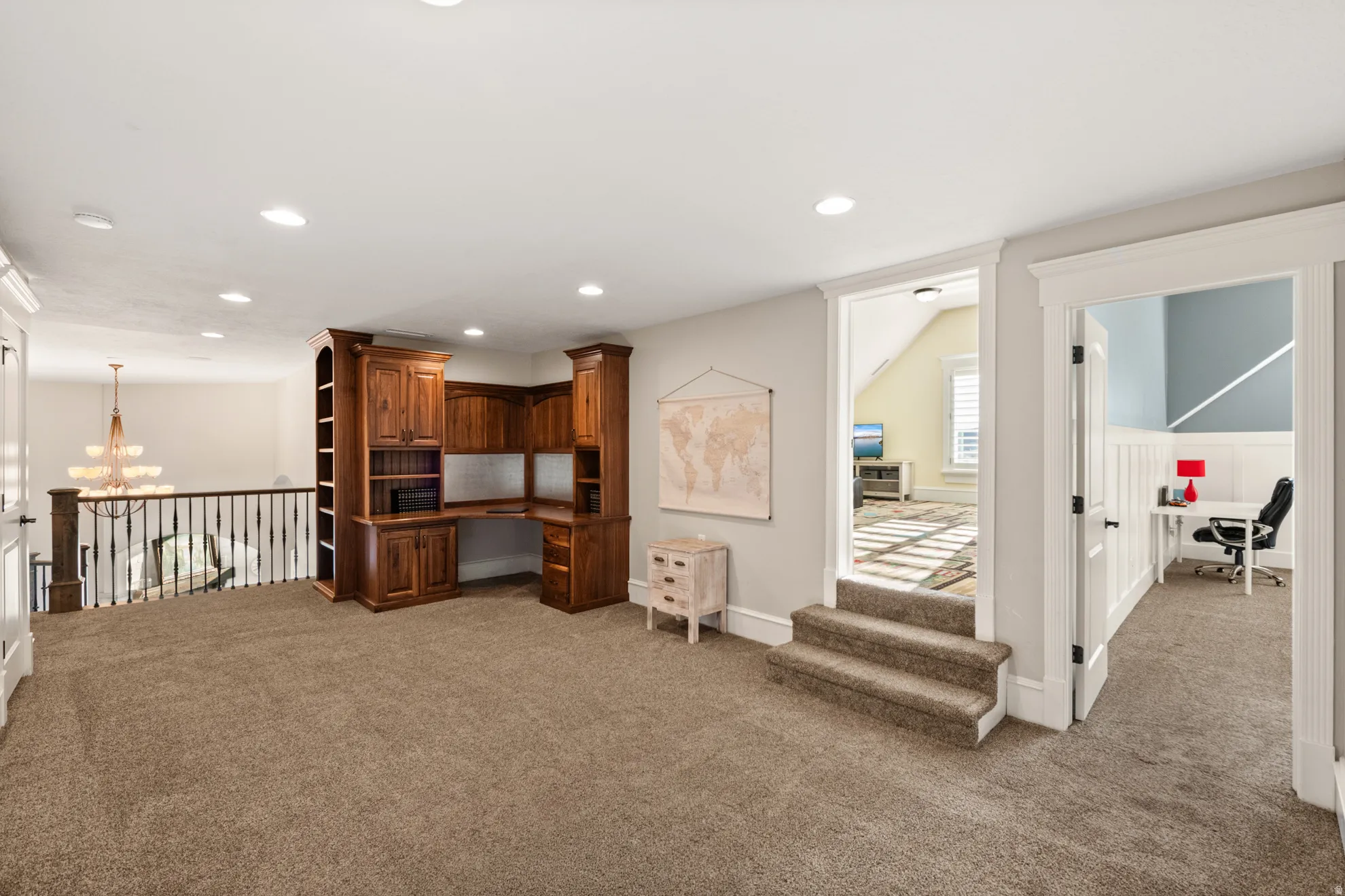 Expansive carpeted loft area featuring a built-in wood-finish desk with integrated shelving, recessed lighting, and a wrought iron banister overlooking the floor below