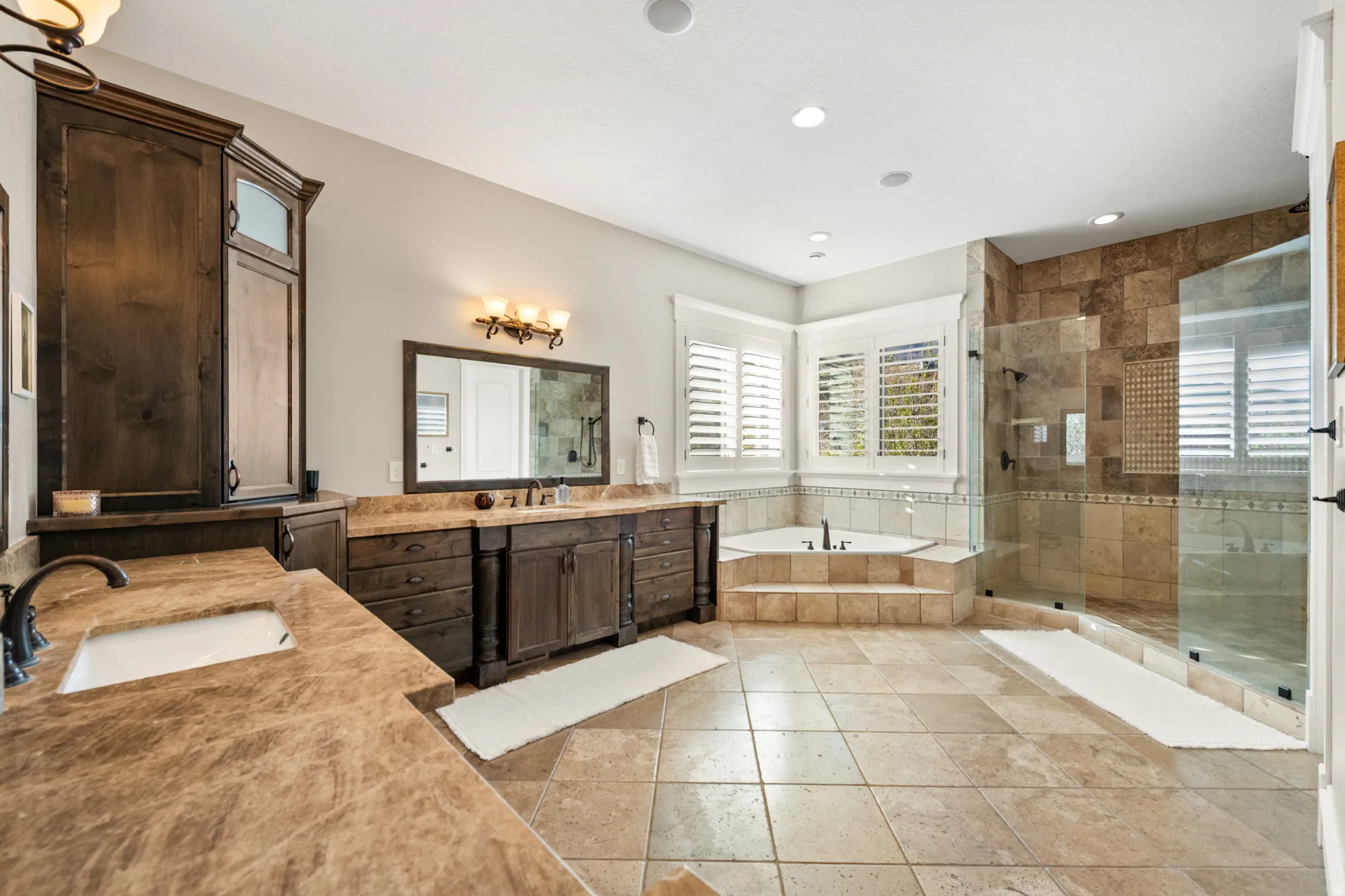 Spacious bathroom featuring natural stone flooring, a corner soaking tub with stepped surround, a glass-enclosed shower with natural stone tile, and dark wood vanities with integrated sinks and stone countertops