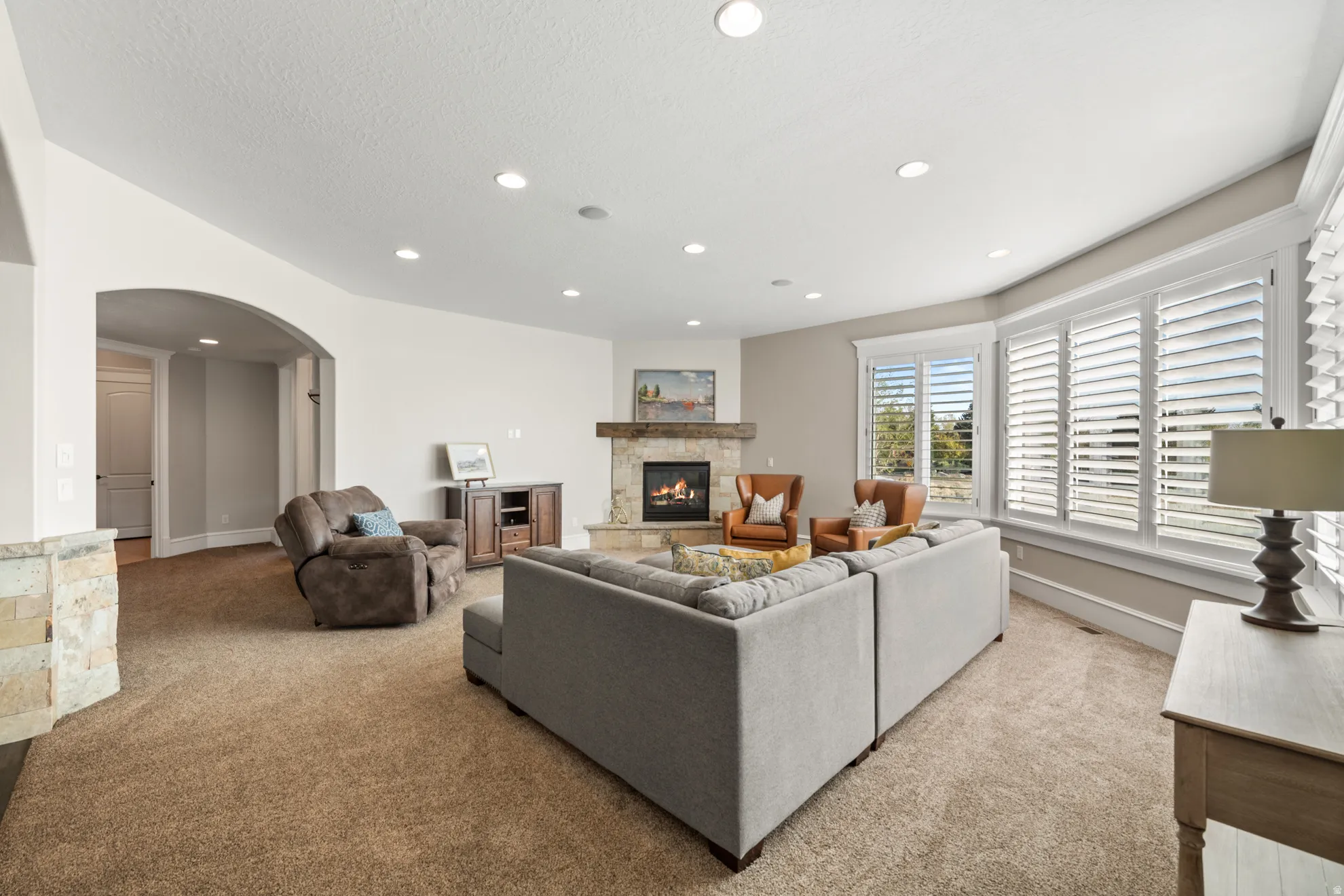 Spacious living area featuring a stone-clad fireplace with a wood mantel, a series of large windows with white plantation shutters, an arched doorway, recessed lighting, and a neutral carpet