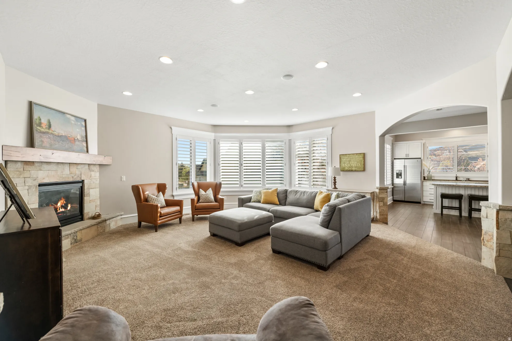 Carpeted living area featuring a stone-clad fireplace with a wood mantel, recessed lighting, and bay windows with plantation shutters