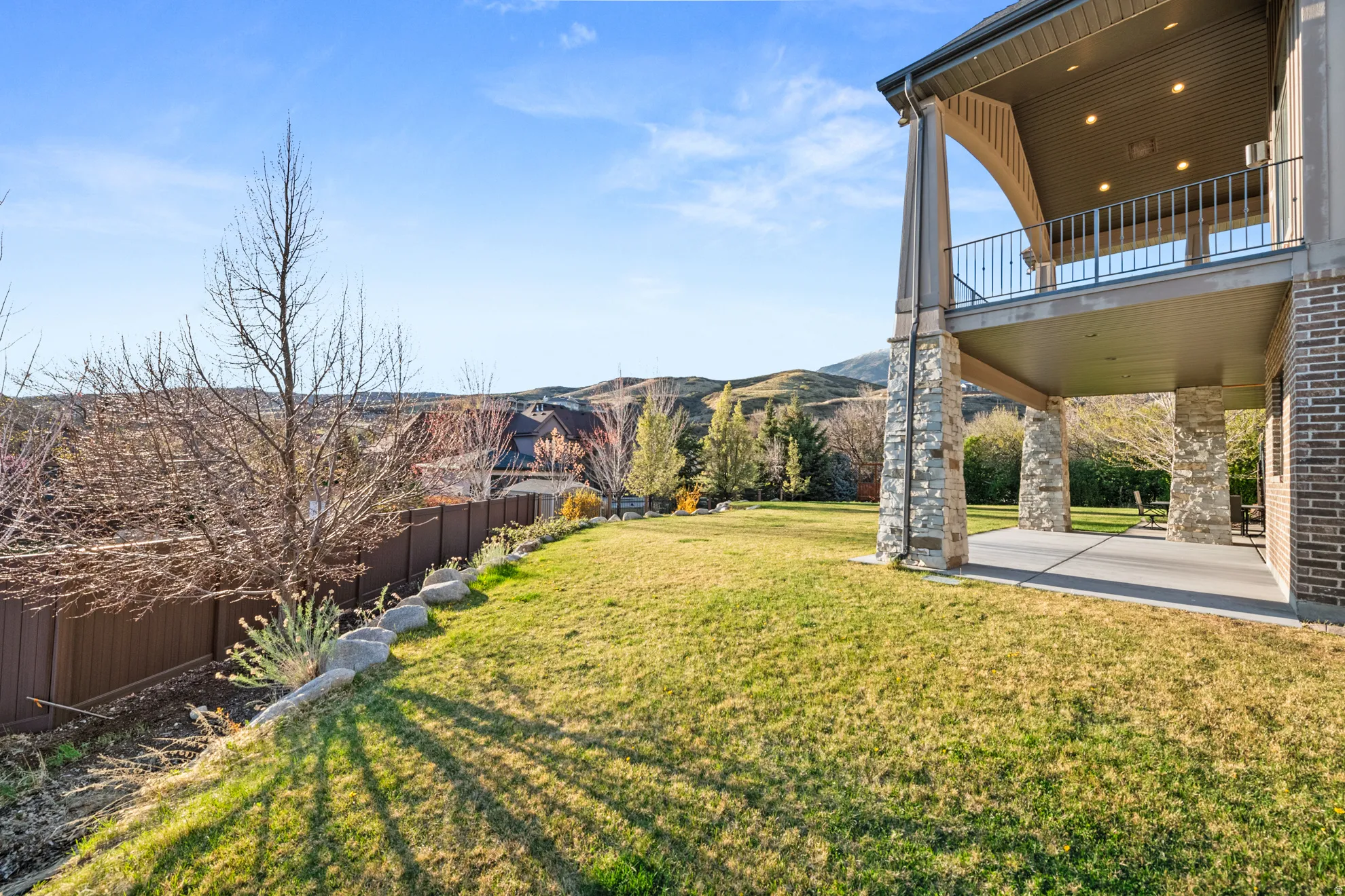 Expansive backyard featuring a manicured lawn, brown privacy fencing, and a stone-clad covered patio