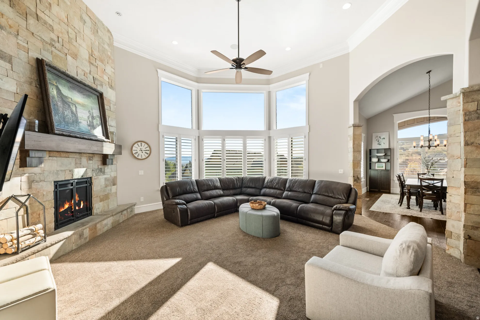 Spacious living area featuring a stone-clad fireplace with wood mantle, large bay windows with plantation shutters, vaulted ceilings, and recessed lighting